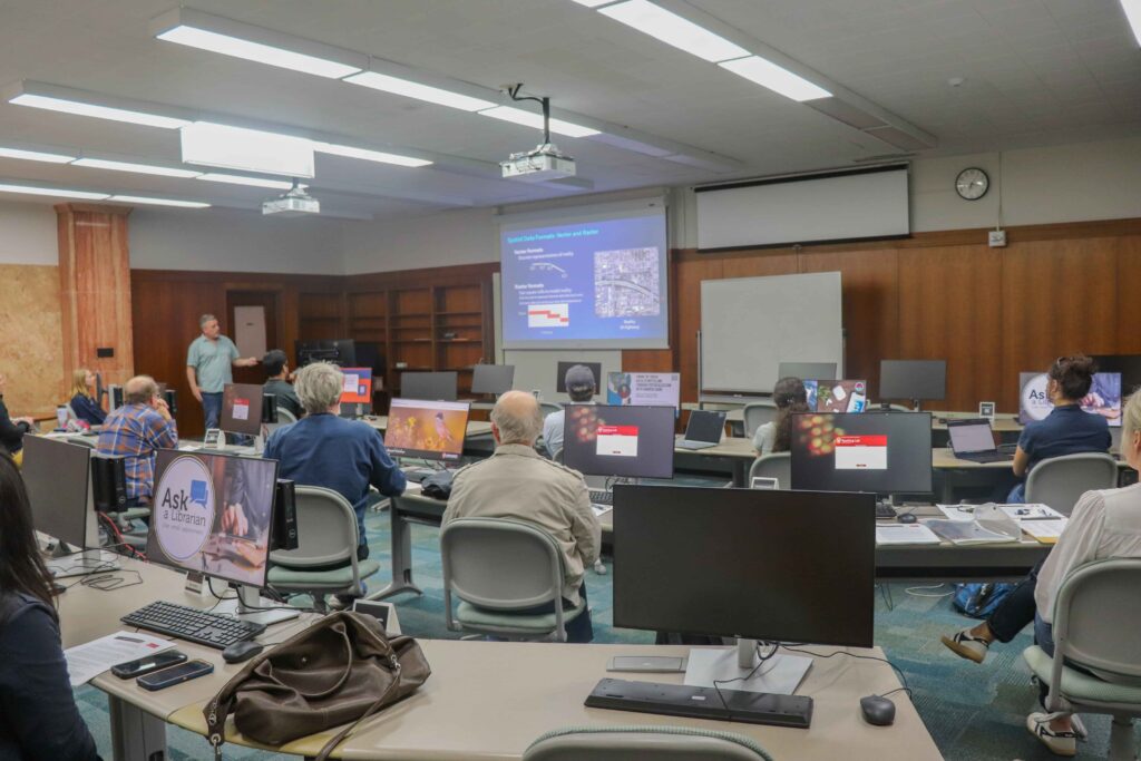 A man at the front of a room points to a screen, while people sitting at three rows of computers watch and listen to him.
