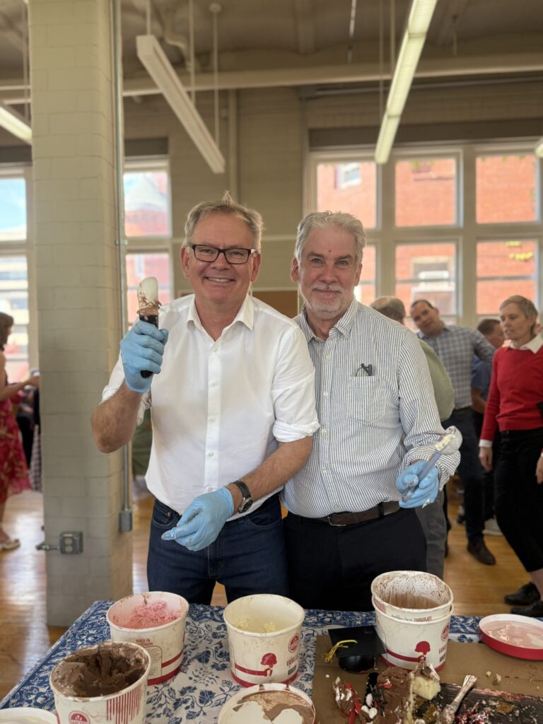 A man in a white shirt holds an ice cream scoop and a man in a striped shirt holds a serving utensil