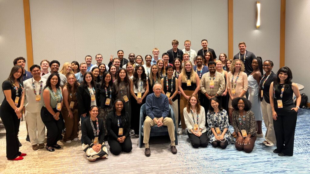 Jack Dangermond sits in a chair; behind him, a large group of students poses for the camera.