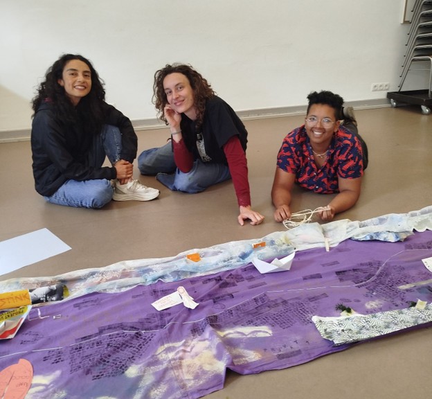 In the foreground, a purple piece of fabric with various markings on it stretches across the floor; in the background, Natalie Correa (right) and two of her teammates pose with smiles.